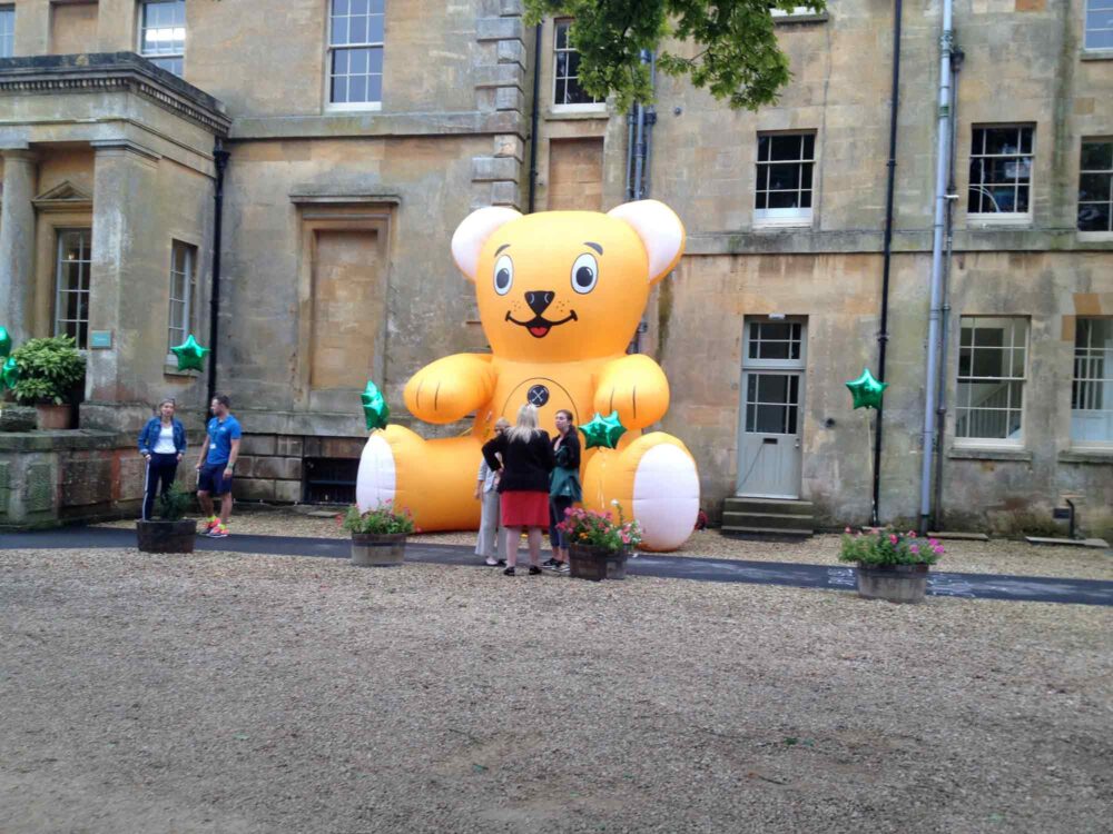 Giant inflatable yellow bear by large house with people in front.